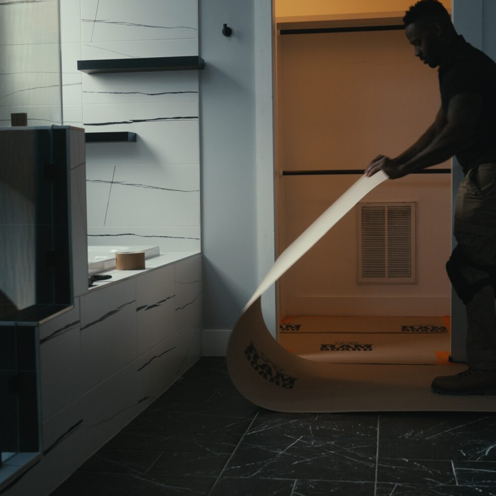 A man in a dark room is unrolling a long, curved beige adhesive strip or mat with black printed text,