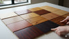 A collection of various hardwood flooring samples arranged on a table, showcasing different wood species, colors, and grain patterns. A person's hands are pointing at two of the samples, highlighting the diversity of wood flooring options.