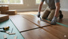 A person is installing glueless click engineered hardwood flooring, showing the process of laying wood planks over an underlayment with tools like a hammer and measuring tape nearby.