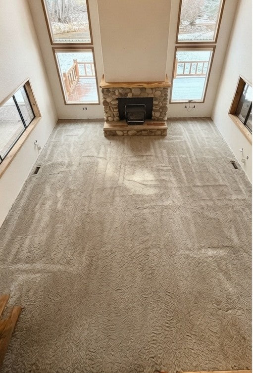 A vaulted living room with a stone fireplace and tall windows, floored in worn beige carpet