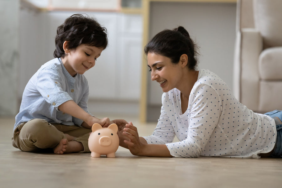 A parent and young child putting coins into a piggy bank together on a wood floor