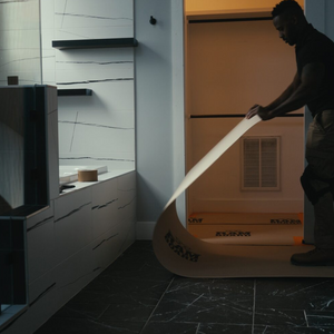 A man in a dark room is unrolling a long, curved beige adhesive strip or mat with black printed text,