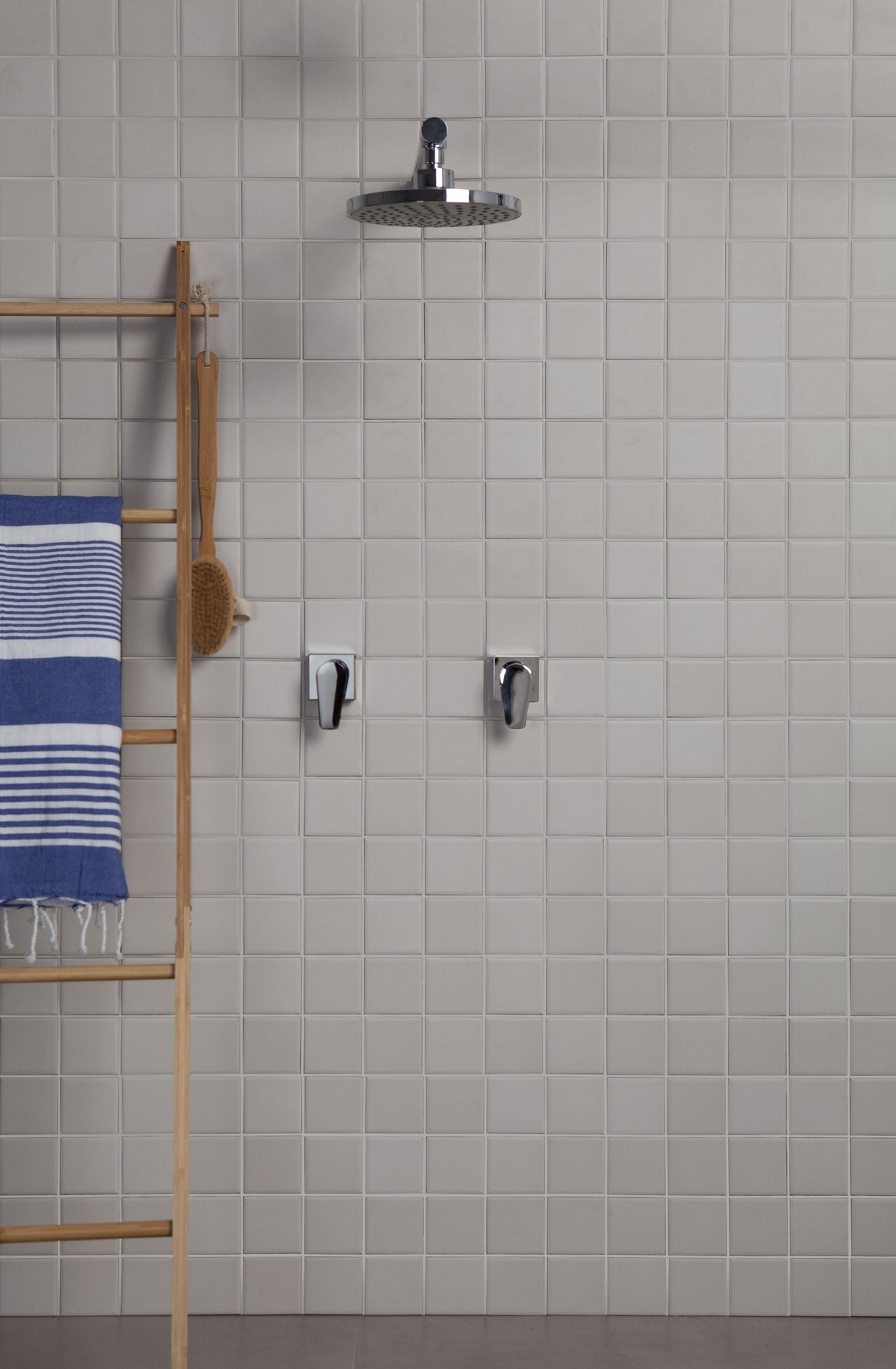 A modern wall-mounted rain shower head with chrome fixtures installed on a light gray tiled bathroom wall, with a wooden