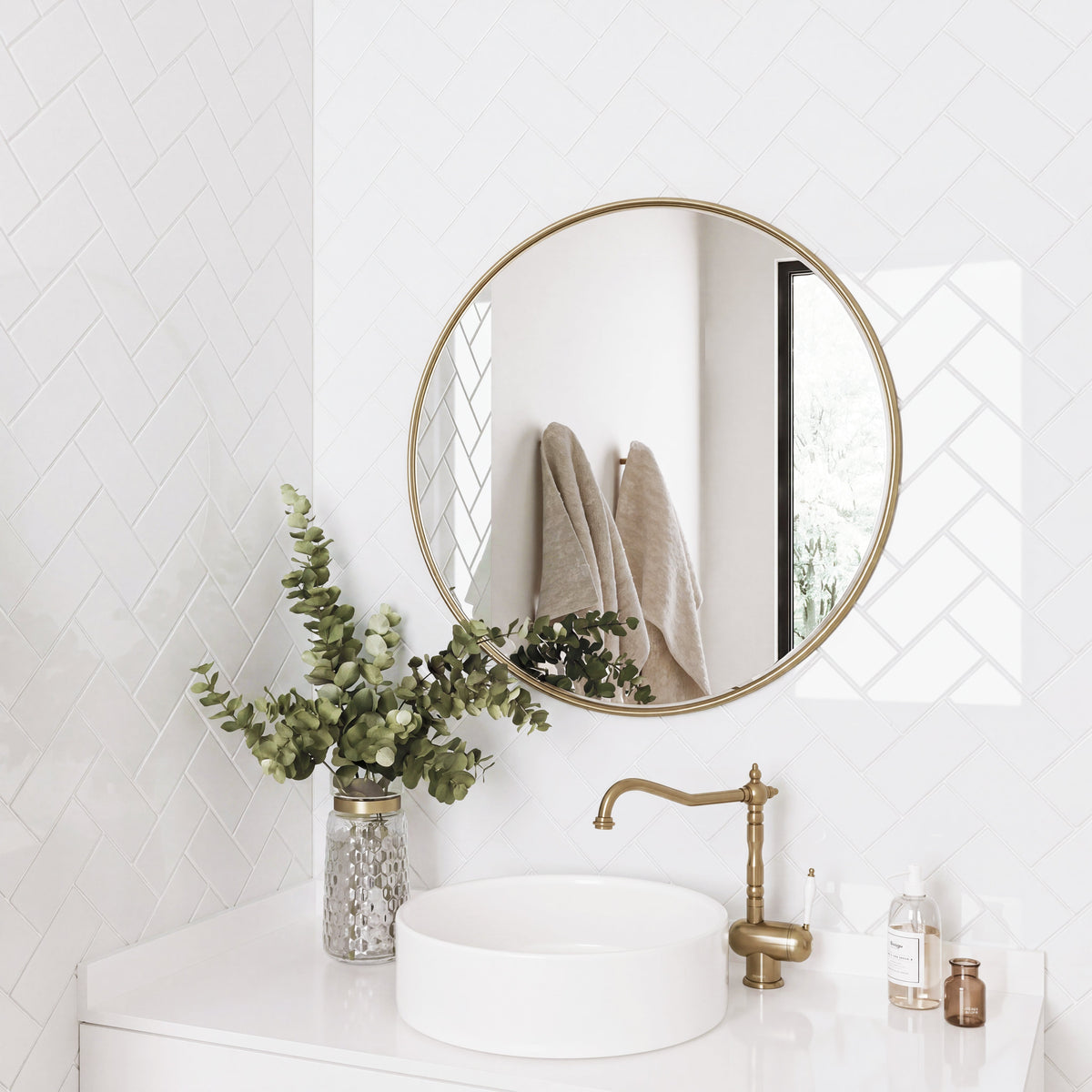 Bathroom with round gold mirror, white subway tiles, white sink, and greenery.